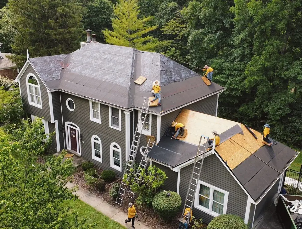 Drone view of a crew installing a new roof on a gray colonial home, with ladders on two sides